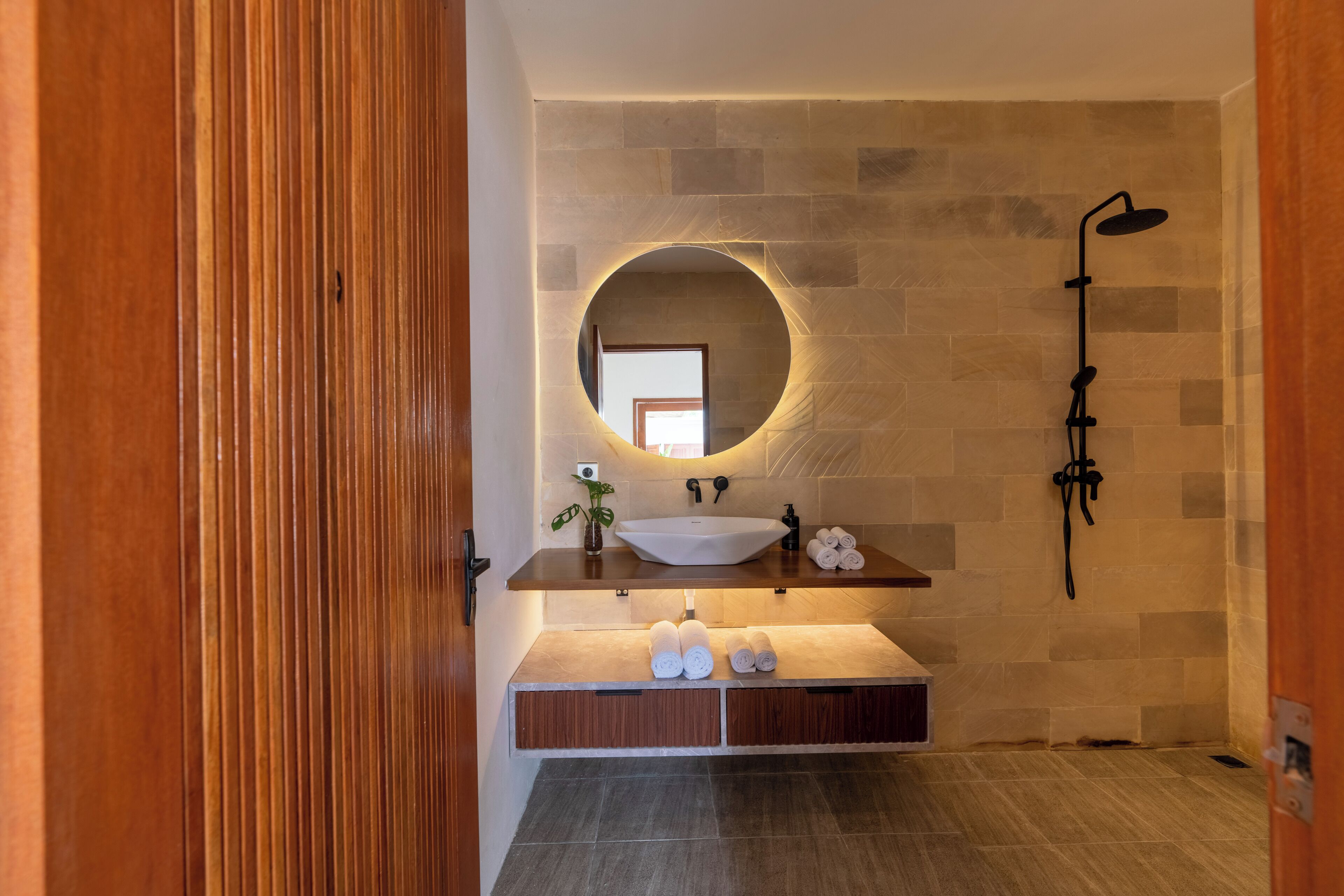 Une salle de bain moderne avec un meuble-lavabo en bois, un miroir rond, un lavabo blanc et un espace douche.