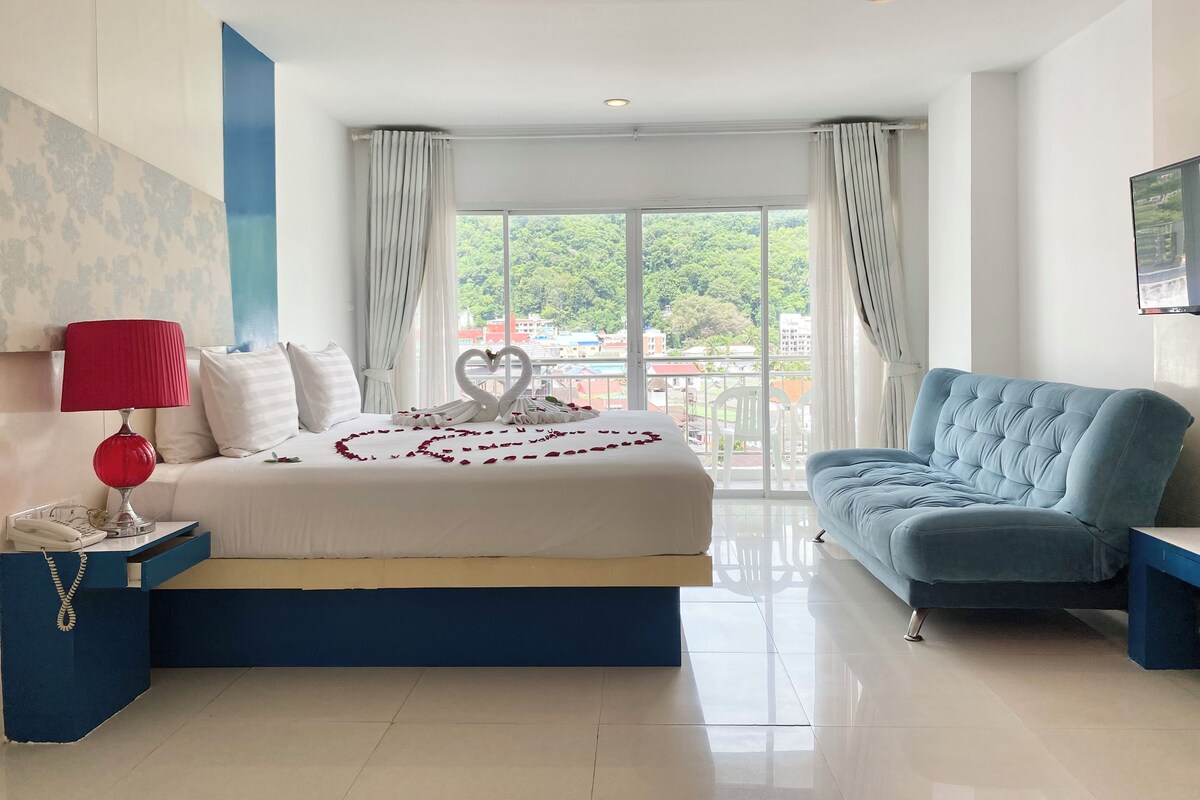 A modern hotel room with a large bed, a blue armchair, a desk with a telephone, and a scenic view through the window.