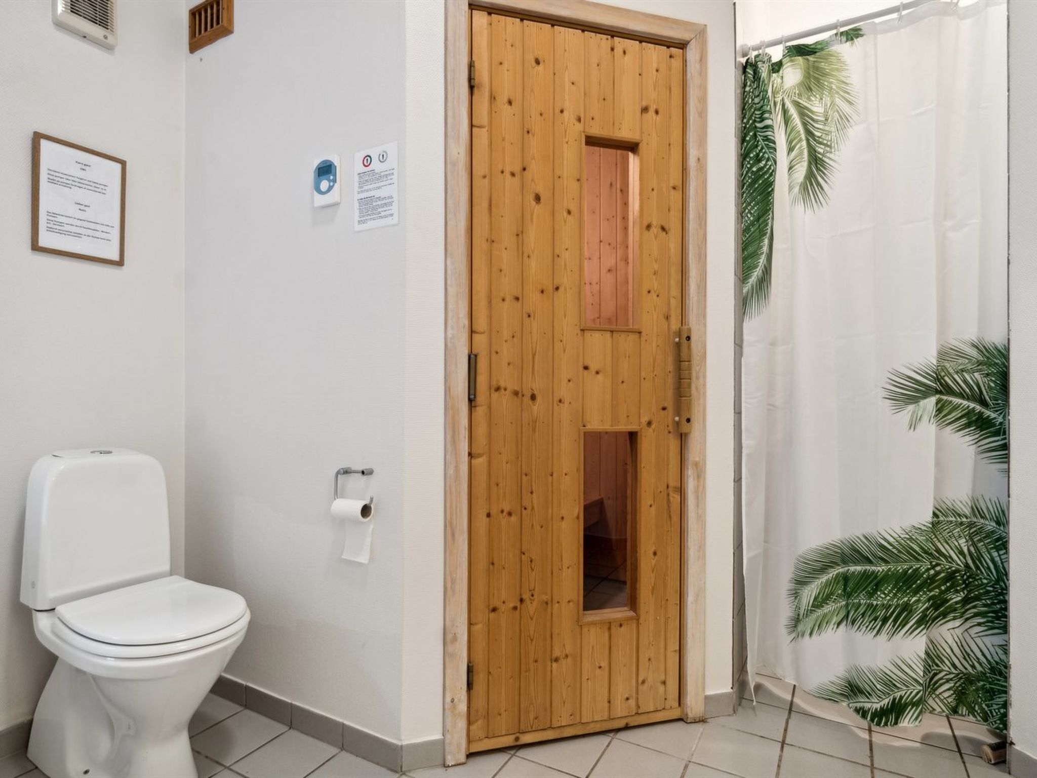 A bathroom with a wooden door, a white toilet, a paper towel dispenser, and a shower curtain with palm leaves.