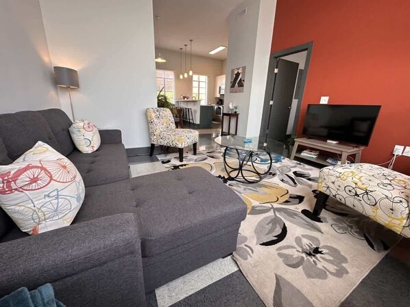 Un salon avec un canapé, un pouf et un tapis à motifs. On y trouve également une télévision et une table avec une lampe.