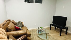 Ein Wohnzimmer mit einem braunen Ledersofa, einem Glastisch, einer schwarzen TV-Konsole und einer kleinen Pflanze auf dem Tisch.