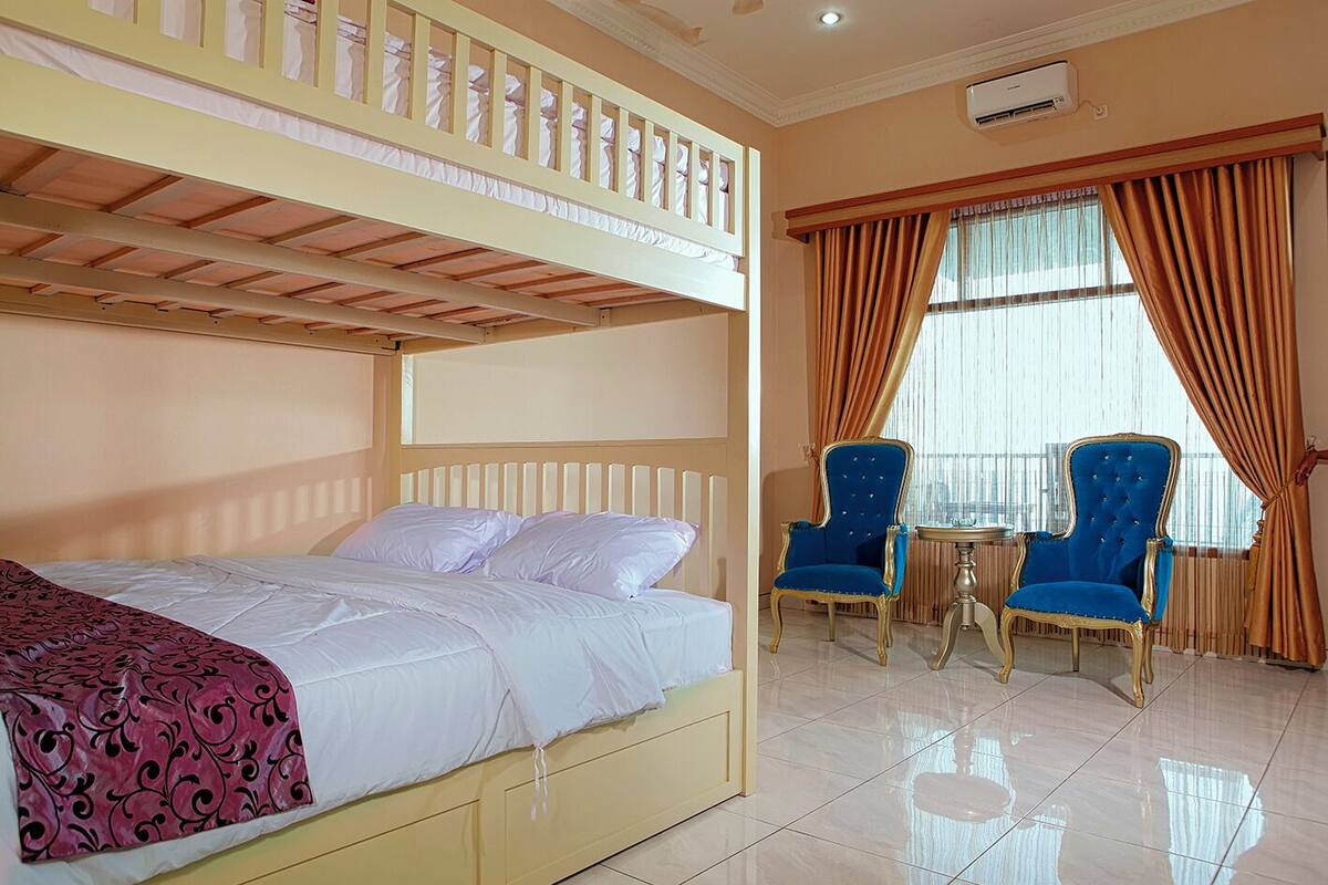 A hotel room with a bunk bed, two blue armchairs, a small table, and a large window with curtains.