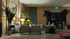 A modern lobby with a sofa set, a red ottoman, and a blue sofa. There are potted plants, a lamp, and a door with an exit sign. - Sugardunes Resort (Al Khaluf)