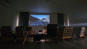 A dark room with a single screen displaying a snowy landscape. There are four wooden chairs and a small table in front of the screen. - Sugardunes Resort (Al Khaluf)