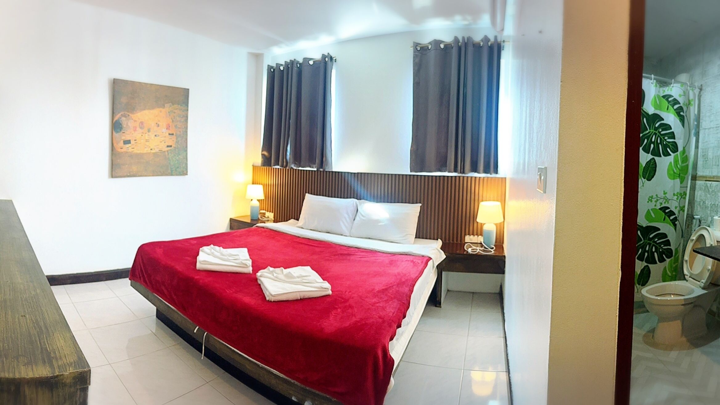 A hotel room with a bed, two bedside tables with lamps, a painting on the wall, and a bathroom visible through an open door.