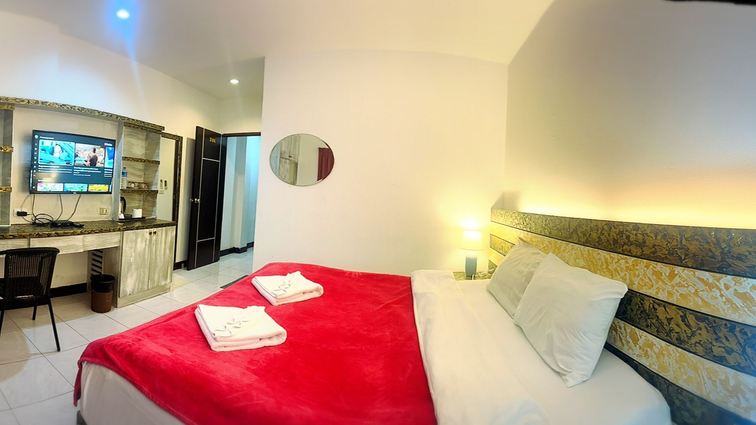 A hotel room with a bed, a desk with a computer, a mirror, and a TV displaying a news channel.