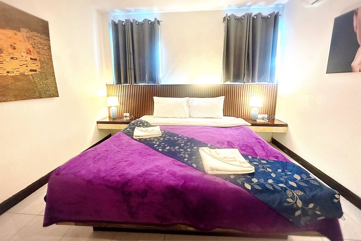 Une chambre d’hôtel avec un lit, deux lampes de chevet, une table de nuit, une fenêtre avec des rideaux et un tableau au mur.