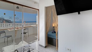 Ein Balkon mit Glastür, ein Bett mit blauer Bettdecke und einem an der Wand befestigten Fernseher.