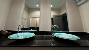 Un baño con dos lavamanos, cada uno con una pileta de cerámica azul y grifos de cromo. Hay un espejo grande sobre los lavamanos, y una unidad de aire acondicionado montada en la pared a la derecha.