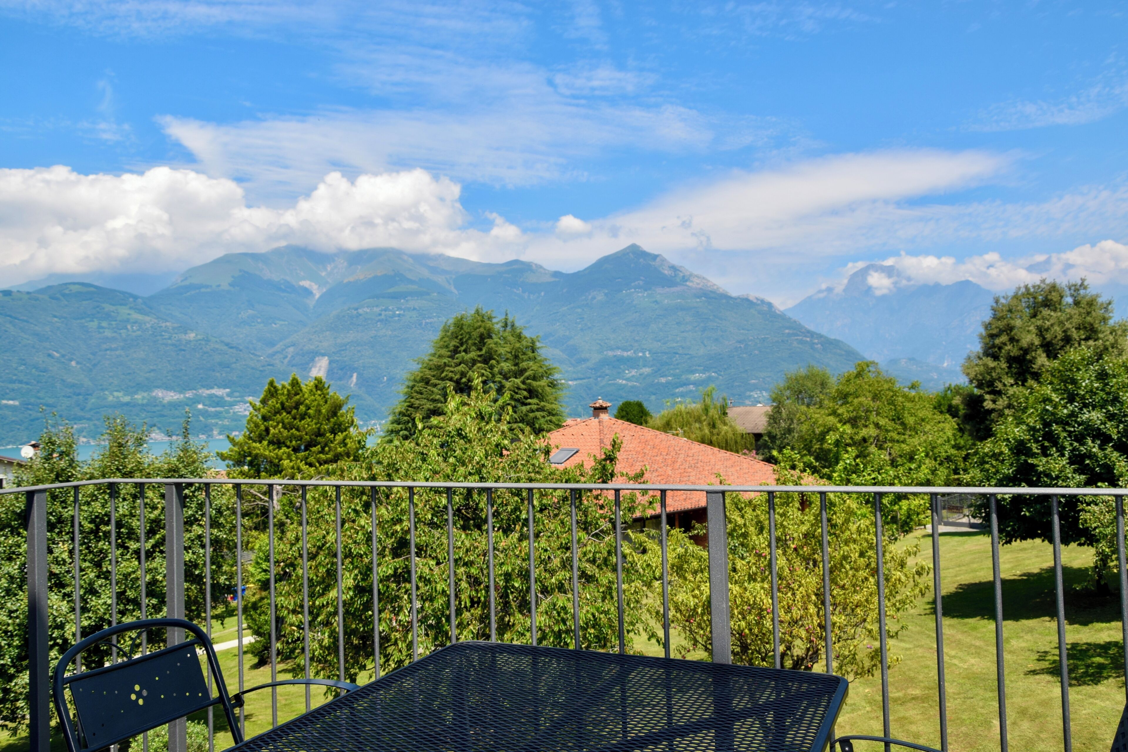Un balcone con ringhiera in metallo che si affaccia su un paesaggio montuoso con alberi e un edificio con tetto rosso.