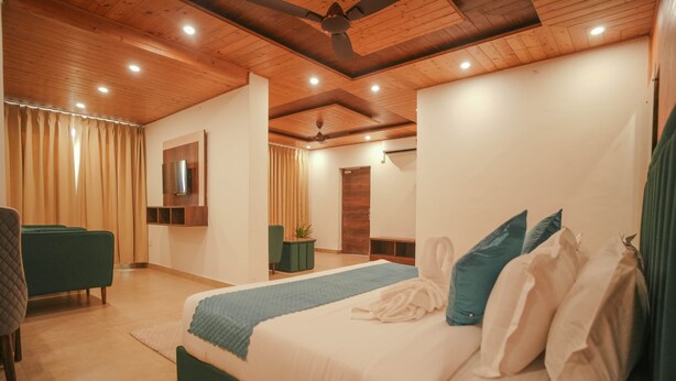 A modern bedroom with a large bed, a green armchair, a TV mounted on the wall, and a ceiling fan. - Tapovan Resorts (Kasauli)