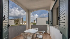 Un balcón con una mesa y dos sillas, vista panorámica de la ciudad y un edificio con contraventanas.