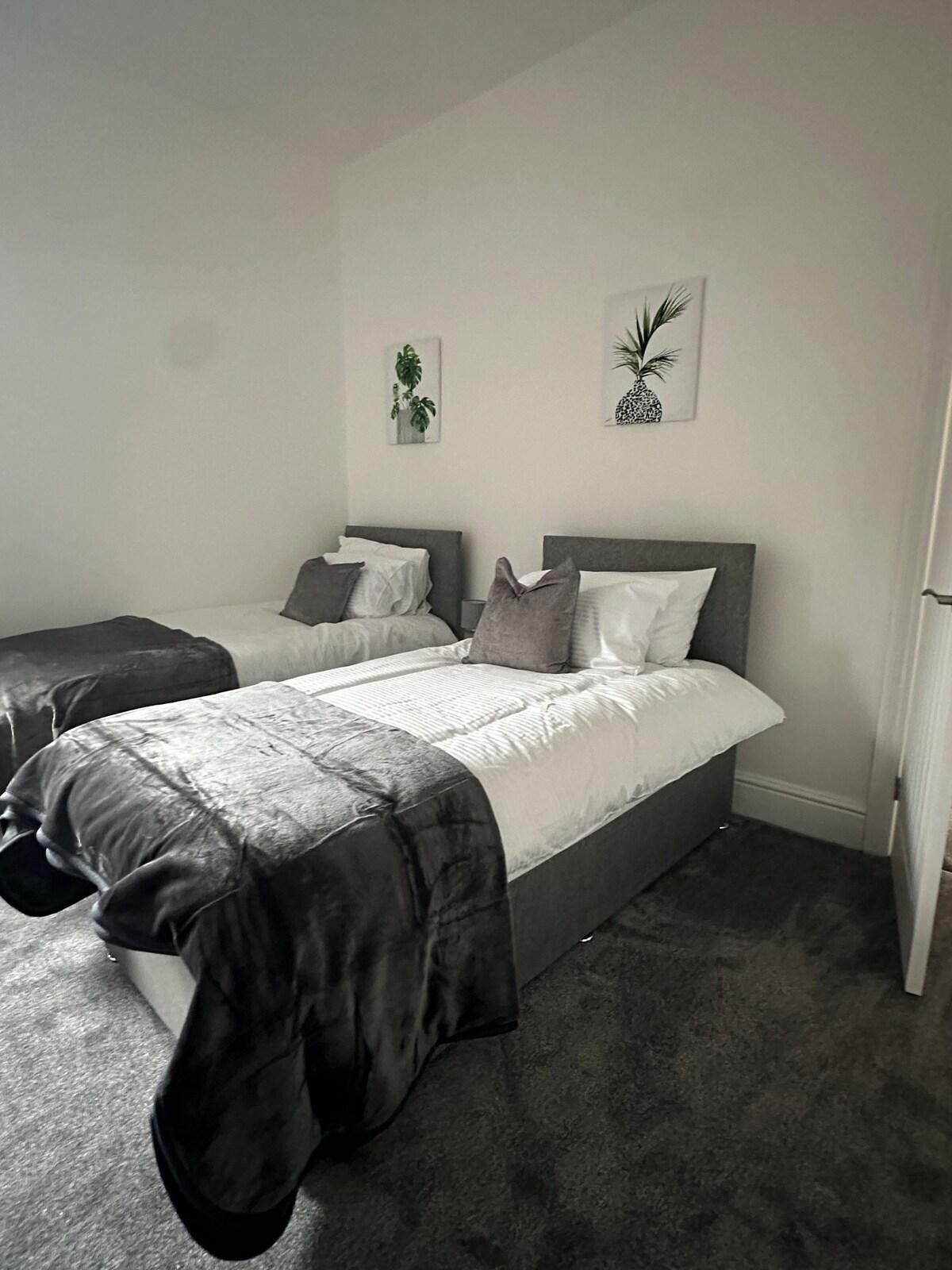 A bedroom with two beds, grey bedding, and two wall-mounted plant pictures.