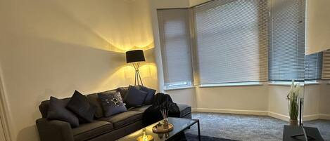 A modern living room with a sofa, a coffee table, a lamp, and a large window with blinds.