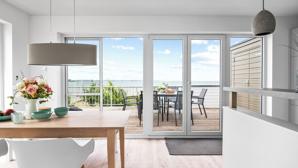A modern dining area with a wooden table, white chairs, and a view of the outdoors through large sliding glass doors.