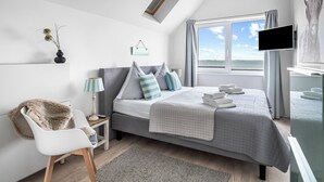 A modern bedroom with a bed, a chair, a bedside table, a lamp, and a window with curtains.