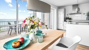 A modern kitchen with a large window overlooking the sea, a dining area with a wooden table, and a white chair.