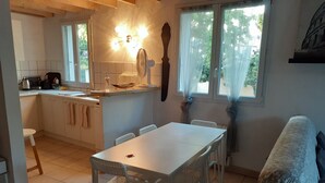 Een compacte keuken met een eethoek, een witte tafel en stoelen. Er is een raam met gordijnen, een spoelbak en een fornuis.