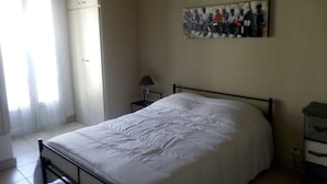 Een slaapkamer met een bed, nachtkastje, lamp, schilderij aan de muur en een raam met gordijnen.