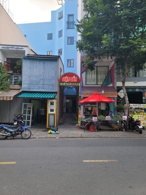 Una escena callejera con un edificio azul, un letrero rojo y blanco para GIAT SAY y un puesto de frutas con etiquetas de precios.