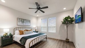 A bedroom with a bed, a nightstand, a lamp, a ceiling fan, a window with blinds, and a potted plant.