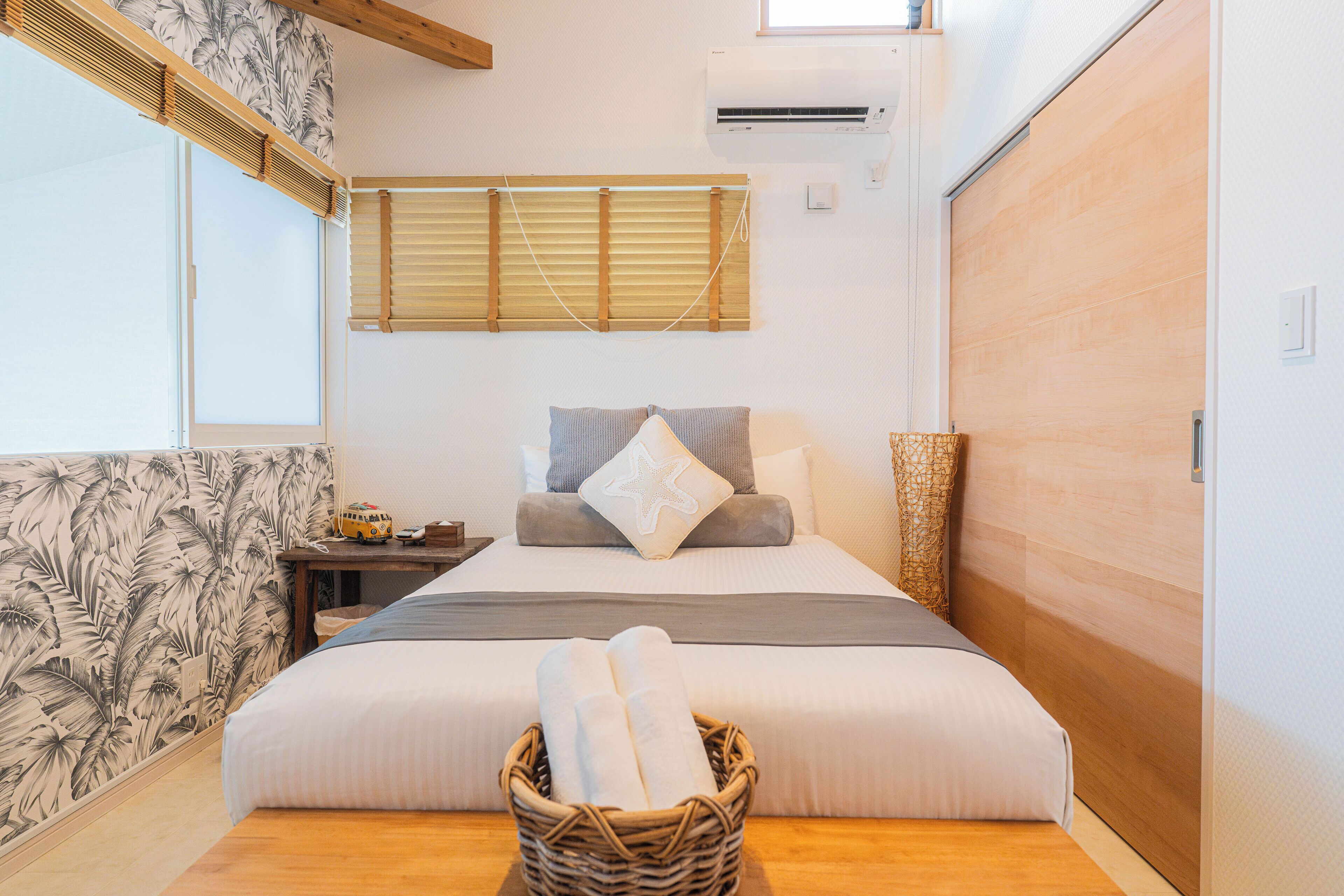 A bedroom with a bed, a nightstand, a wicker basket with towels, a window with blinds, and an air conditioning unit.