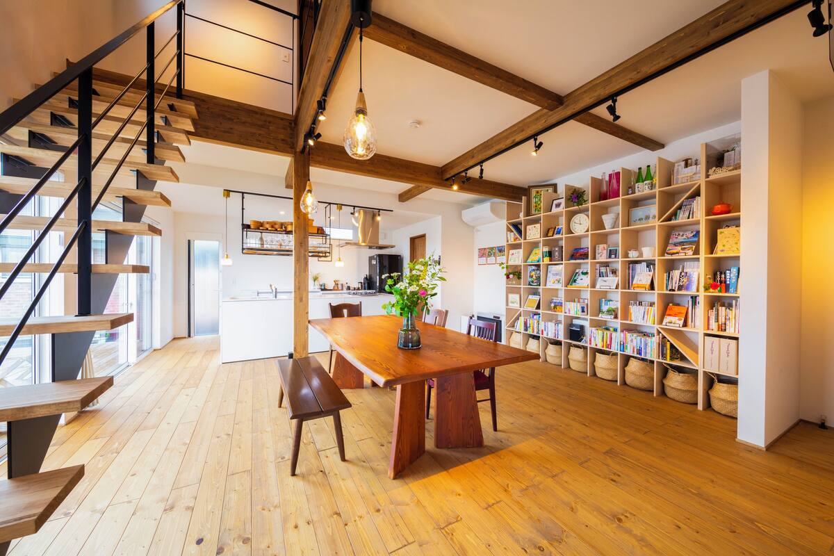 A spacious room with a wooden staircase, a dining table with benches, and a bookshelf filled with books and decorative items.