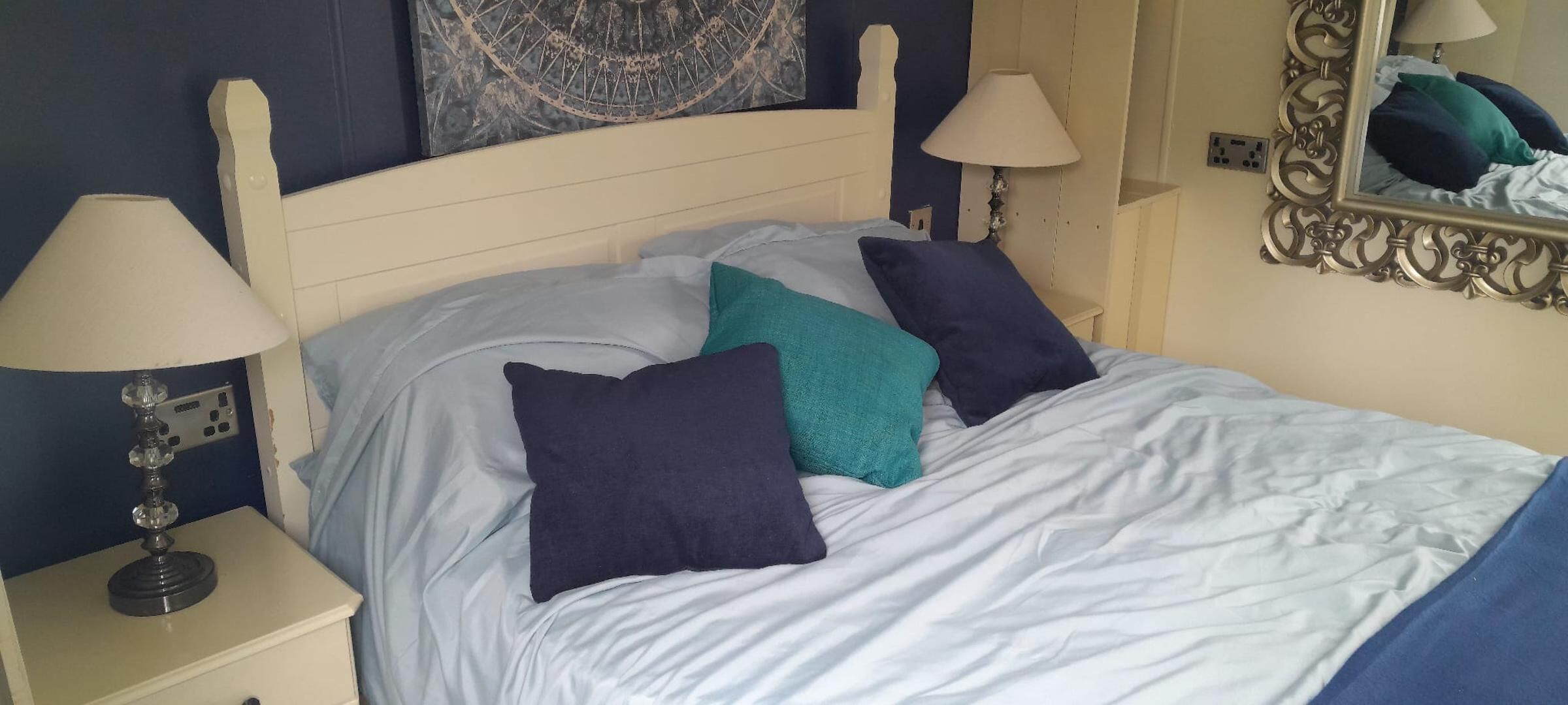 A bed with white linens, teal and dark blue pillows, and a white headboard. There are two bedside lamps and a mirror on the wall.
