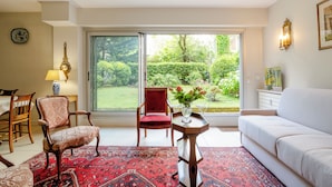 A living room with a red patterned rug, a white sofa, a red chair, a wooden side table with a vase of flowers, and a view of a garden through a glass door.