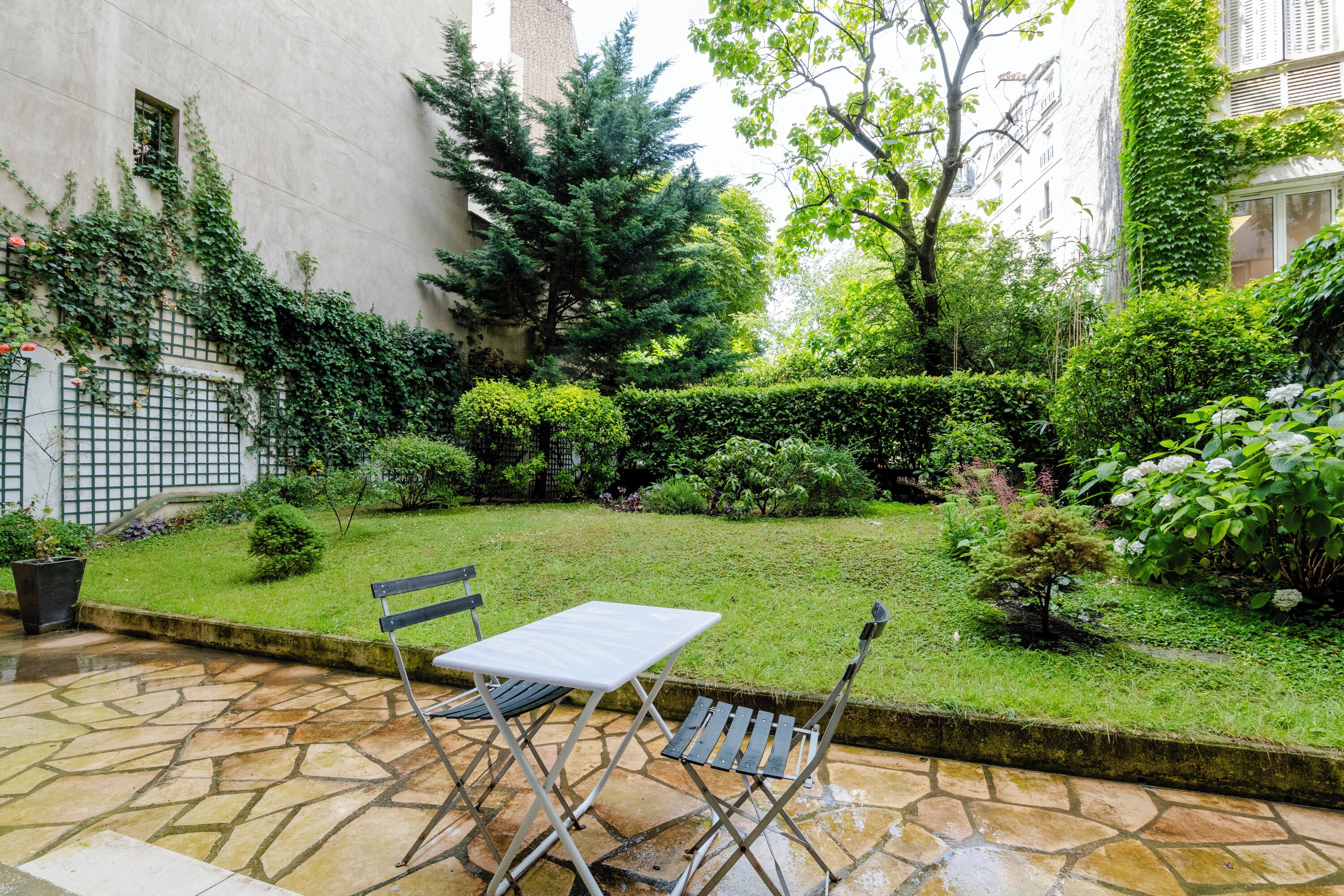 A stone patio with a table and chairs, surrounded by a well-maintained garden with various plants and a hedge.