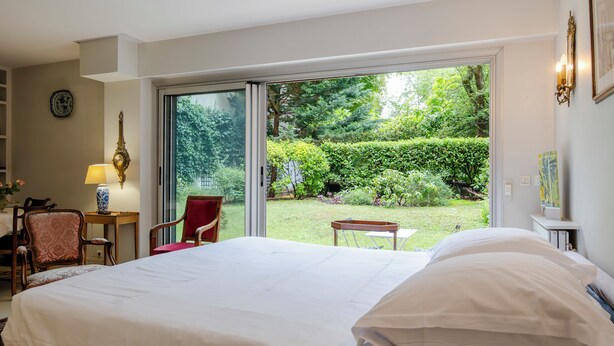 A hotel room with a large bed, a chair, a small table, and a view of a garden through a sliding glass door.