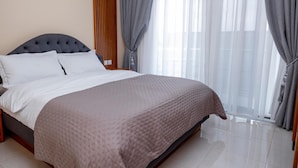 Una cama bien hecha con un cabecero oscuro y almohadas blancas, una colcha color beige y un ventanal con cortinas translúcidas.