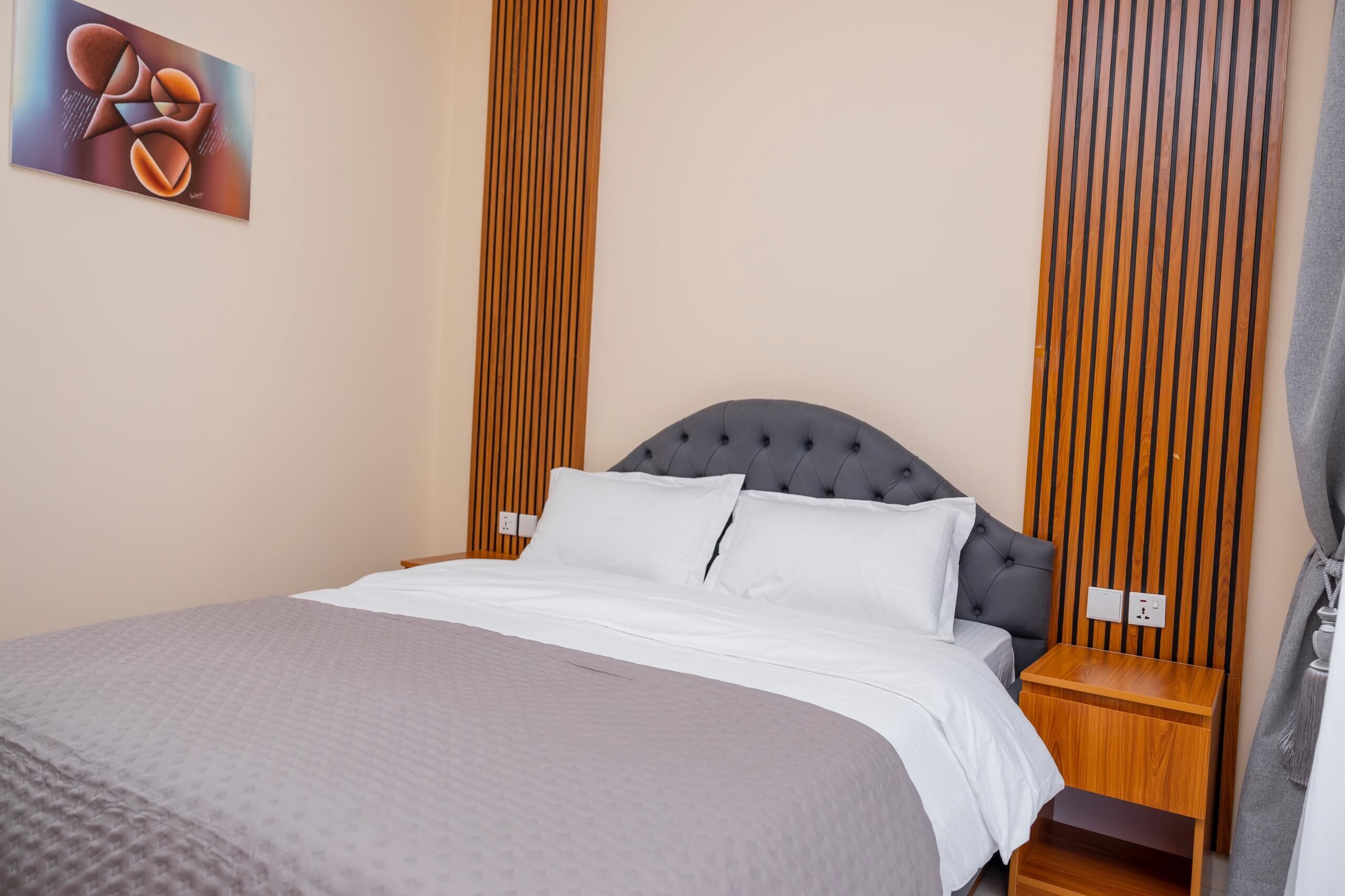 A neatly made bed with white linens, a grey headboard, and a wooden nightstand with a lamp.