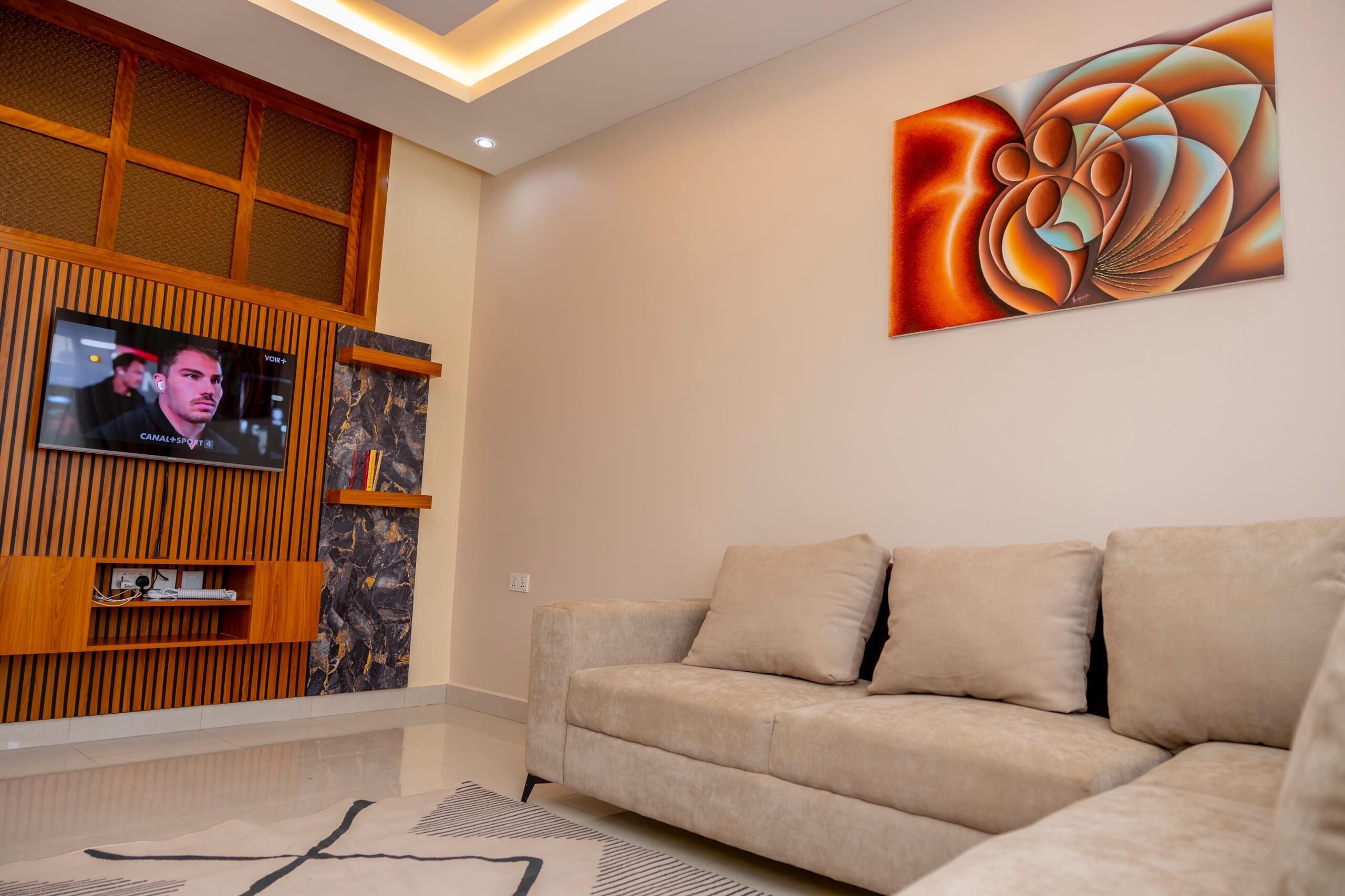 A modern living room with a beige sofa, a mounted TV, and a large abstract painting on the wall.