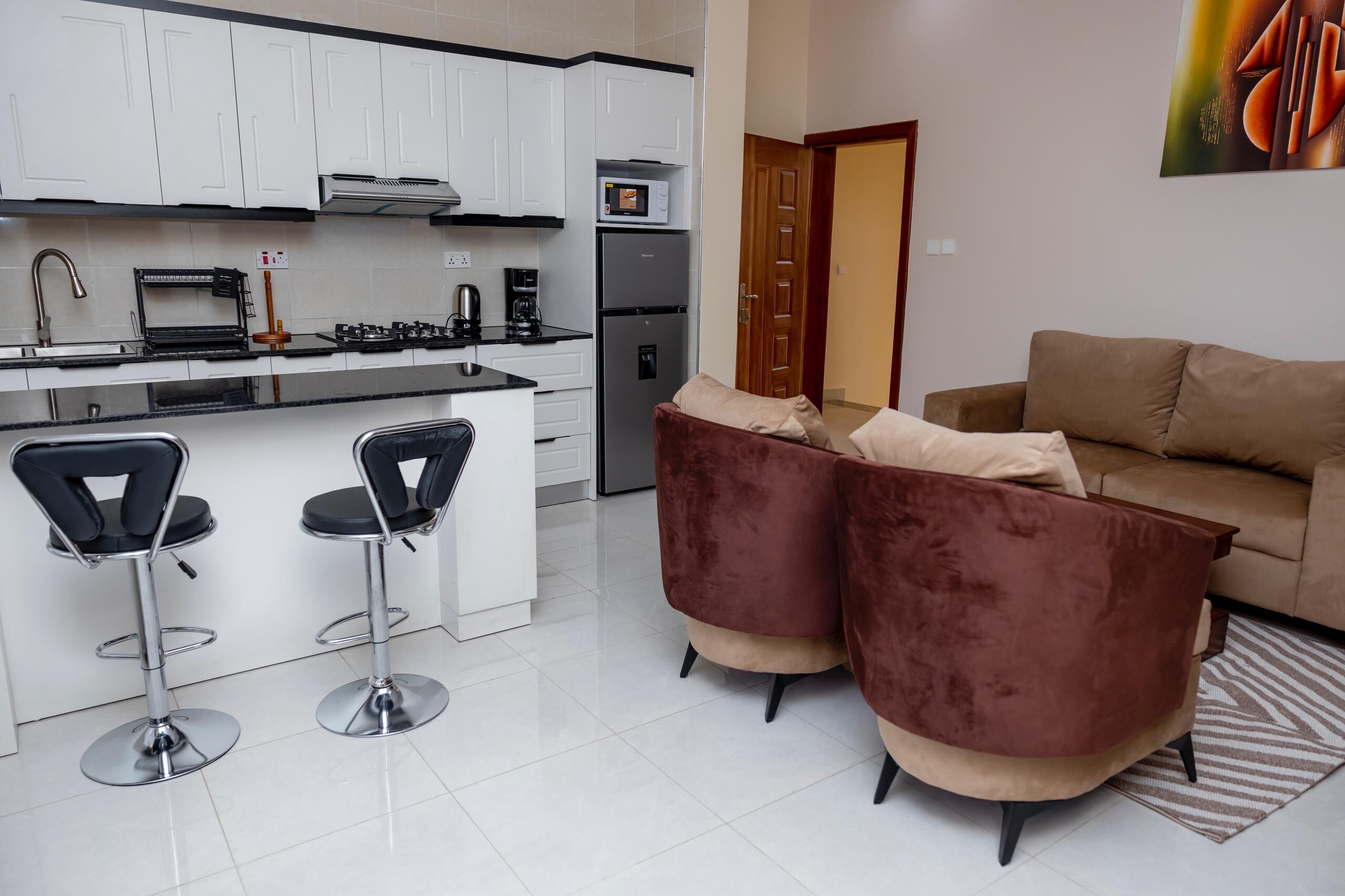 A modern kitchen with white cabinets, a built-in oven, and a refrigerator. There are two bar stools and a brown sofa in the living area.