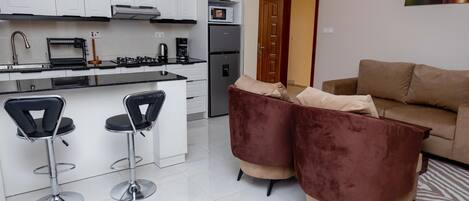A modern kitchen with white cabinets, a built-in oven, and a refrigerator. There are two bar stools and a brown sofa in the living area.