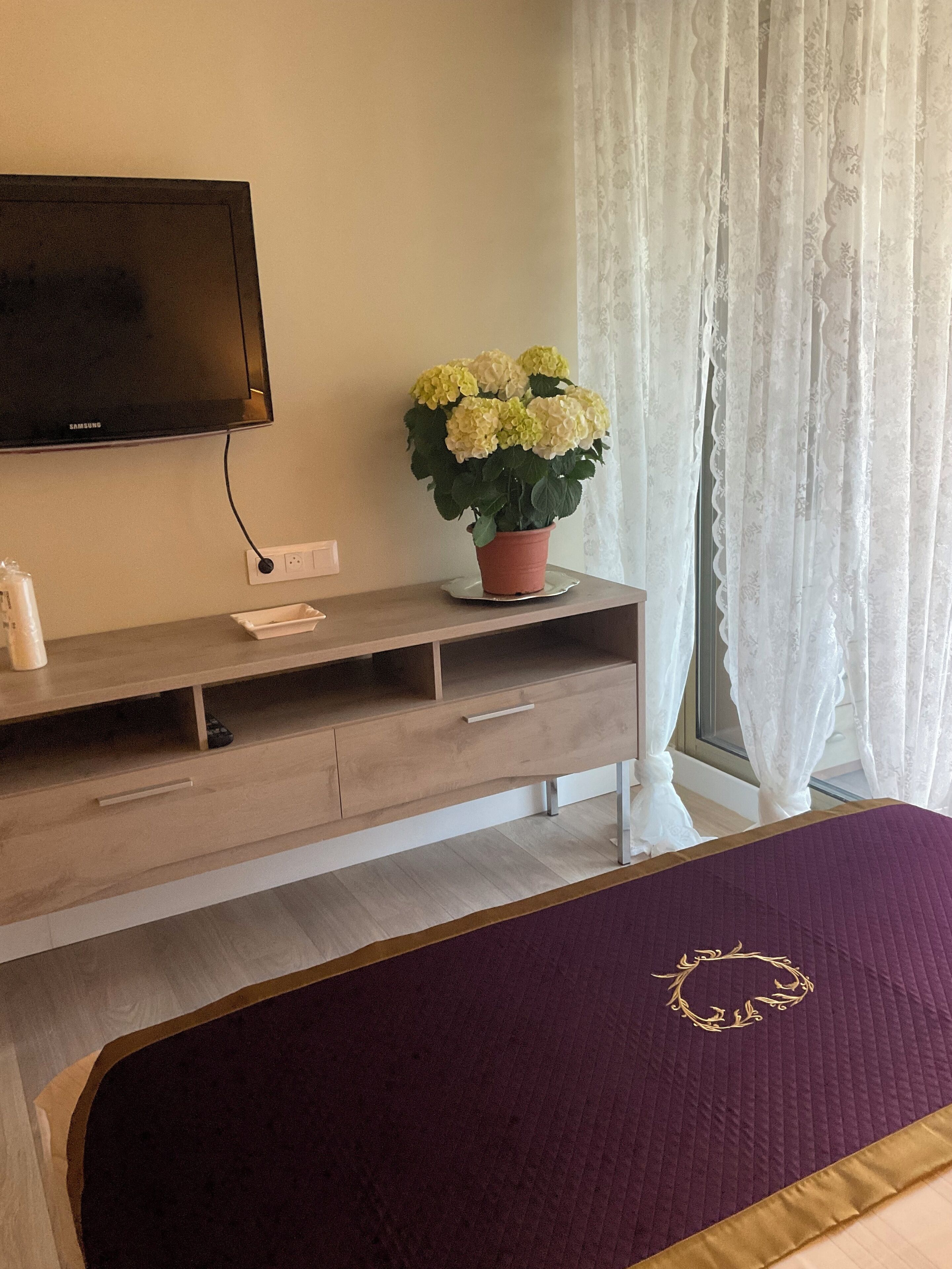 Un dormitorio moderno con una televisión de pantalla plana, una mesa consola de madera con una planta en maceta y una cama con edredón morado.