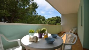 A small round table with chairs on a balcony, surrounded by potted plants and a white chair.