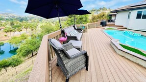 Terraza de madera con mobiliario de mimbre, piscina y vistas a un lago y colinas.