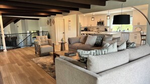 Una sala moderna con sofá, sillón y una mesa de centro. Se ve una cocina al fondo, con un microondas y armarios.