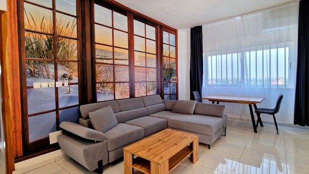 Ein modernes Wohnzimmer mit einem grauen Ecksofa, einem Holztisch und einem Esstisch mit Stühlen. Große Fenster bieten Ausblicke ins Freie.