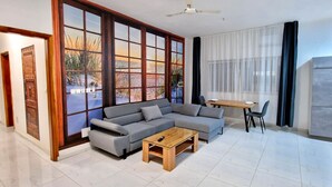 Ein modernes Wohnzimmer mit einem grauen Ecksofa, einem Holztisch und einem Essbereich mit Tisch und Stühlen. Große Fenster bieten einen Blick ins Freie.