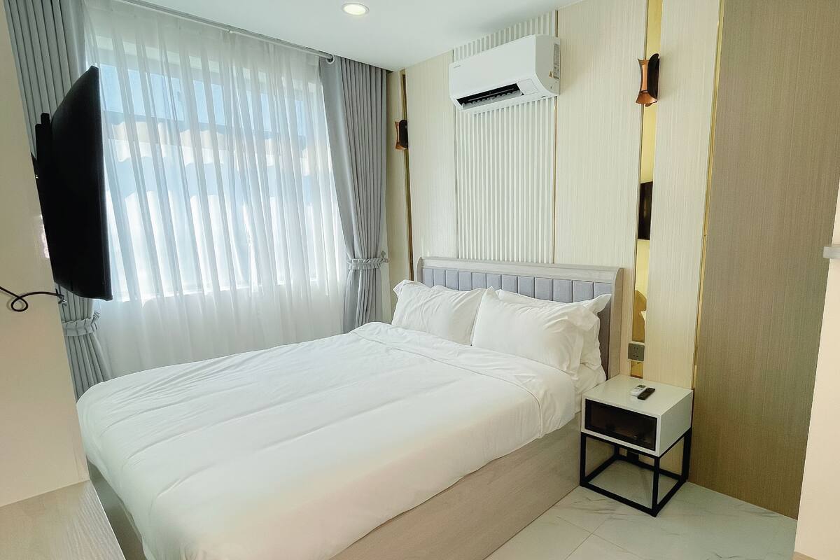 A hotel room with a large bed, a TV mounted on the wall, an air conditioning unit, and a bedside table with a remote control.