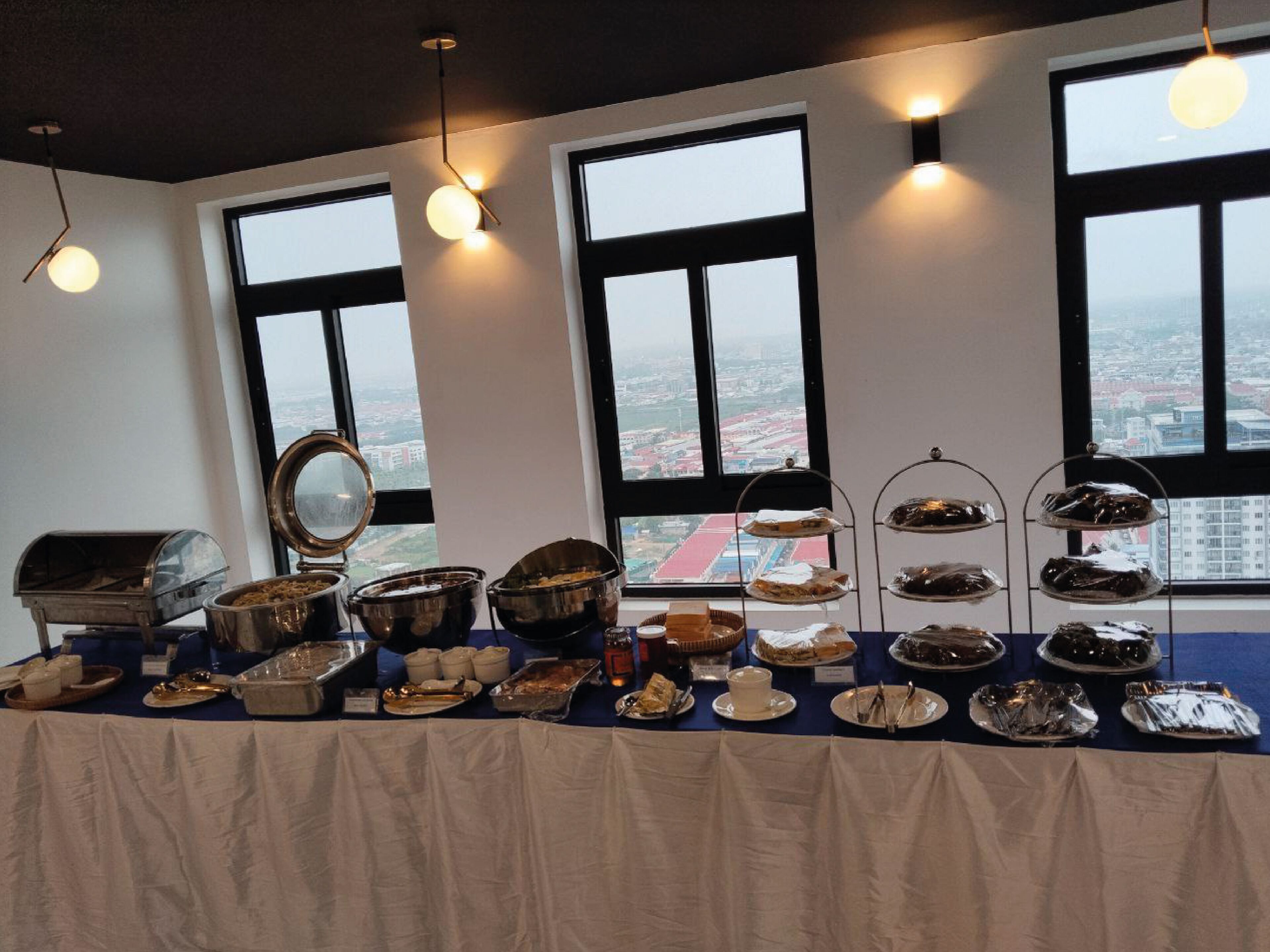A buffet table with various food items and a grill, set against a backdrop of large windows overlooking a cityscape.