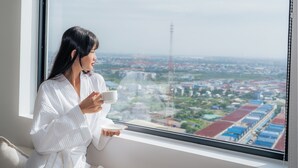 Una persona vestida con una bata blanca sosteniendo una taza y un bol, sentada junto a una ventana con vistas a la ciudad.
