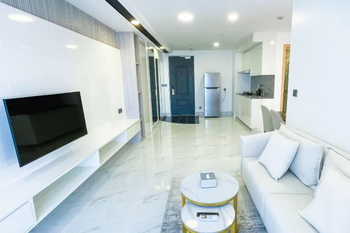 A modern hotel room with a flat-screen TV, a sofa, a small round table, a kitchen area, and a door leading to another room.