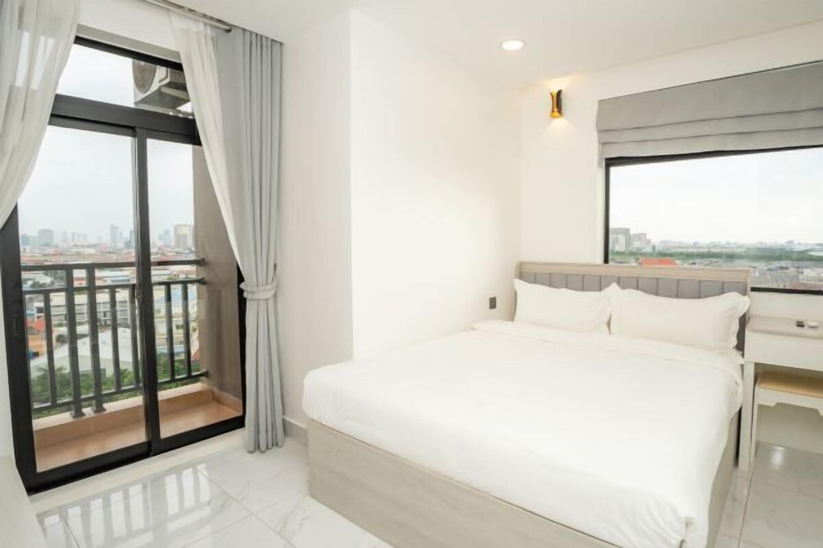 A hotel room with a large bed, a nightstand, a window with a view of the city, and a balcony with curtains.