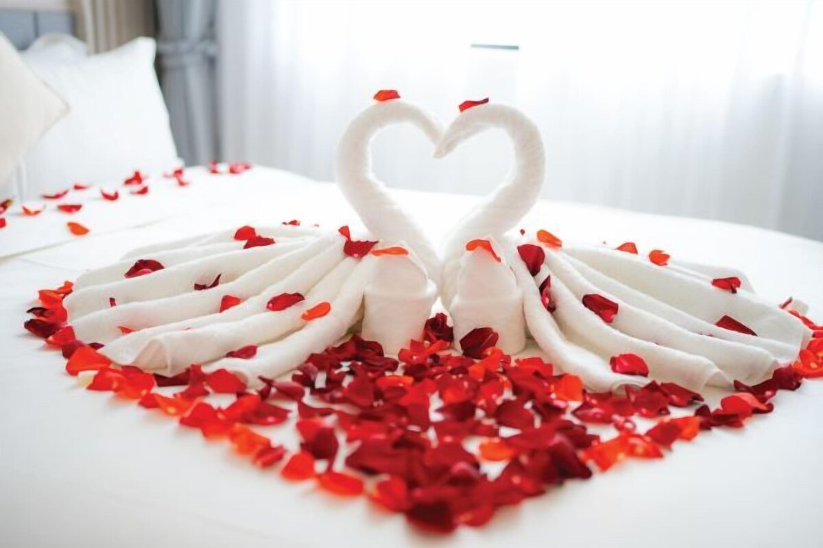 A bed with a white sheet and a heart-shaped arrangement of white towels and red rose petals.