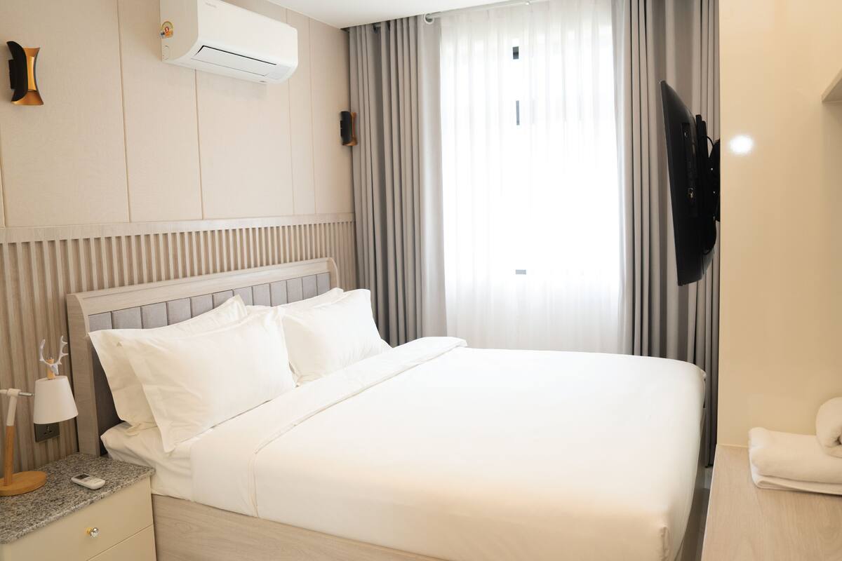 A hotel room with a bed, a TV, a bedside table with a lamp, and a window with curtains.