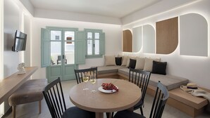 A modern dining area with a round table, chairs, and a bowl of grapes. There is a sofa with cushions and a television mounted on the wall. - Katerina Villas 11 12 13 & 3PriveJacuzzi (Santorini)